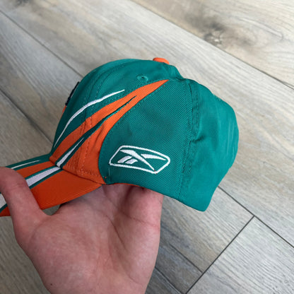 Reebok NFL Miami Dolphins Sideline Baseball Cap is a turquoise baseball cap featuring embroidered Miami Dolphins branding on the front. It includes orange and white accent detailing across the design. The side is finished with a white embroidered Reebok logo, while the back and inside features NFL Equipment tags. The construction uses cotton with a structured fit and a classic adjustable design.