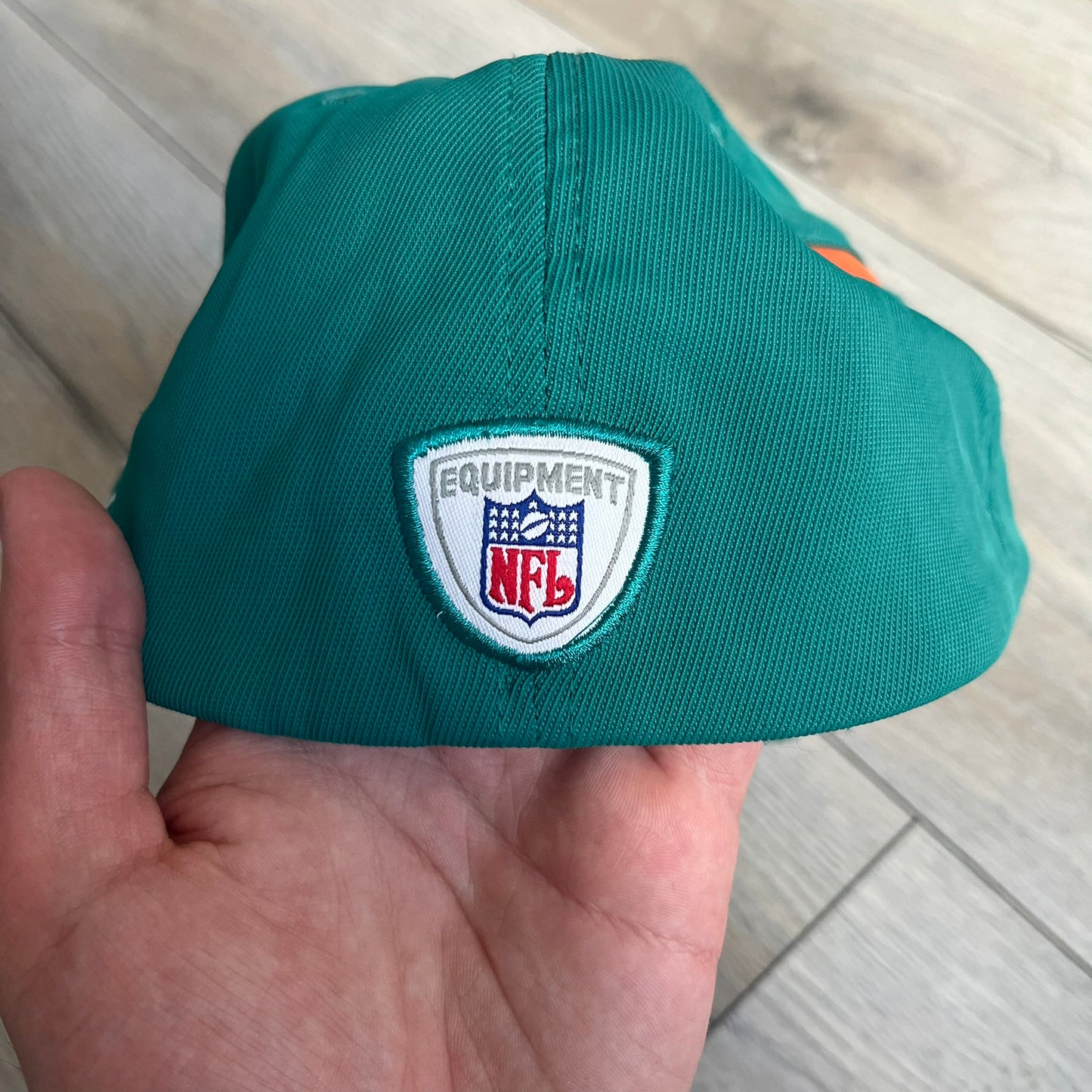 Reebok NFL Miami Dolphins Sideline Baseball Cap is a turquoise baseball cap featuring embroidered Miami Dolphins branding on the front. It includes orange and white accent detailing across the design. The side is finished with a white embroidered Reebok logo, while the back and inside features NFL Equipment tags. The construction uses cotton with a structured fit and a classic adjustable design.
