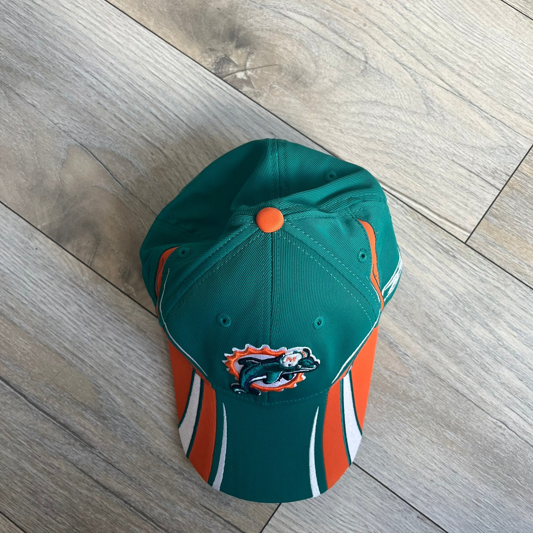 Reebok NFL Miami Dolphins Sideline Baseball Cap is a turquoise baseball cap featuring embroidered Miami Dolphins branding on the front. It includes orange and white accent detailing across the design. The side is finished with a white embroidered Reebok logo, while the back and inside features NFL Equipment tags. The construction uses cotton with a structured fit and a classic adjustable design.