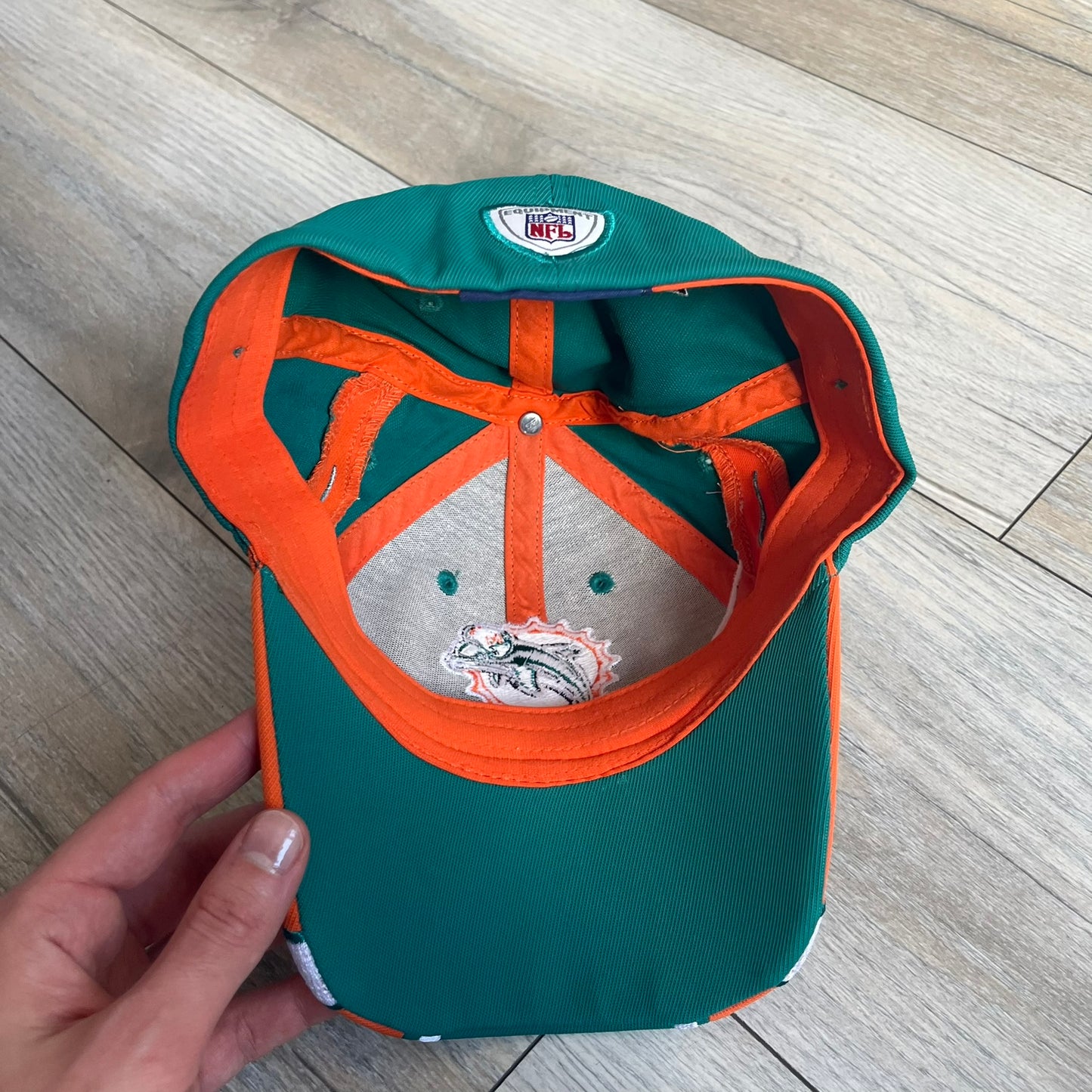 Reebok NFL Miami Dolphins Sideline Baseball Cap is a turquoise baseball cap featuring embroidered Miami Dolphins branding on the front. It includes orange and white accent detailing across the design. The side is finished with a white embroidered Reebok logo, while the back and inside features NFL Equipment tags. The construction uses cotton with a structured fit and a classic adjustable design.