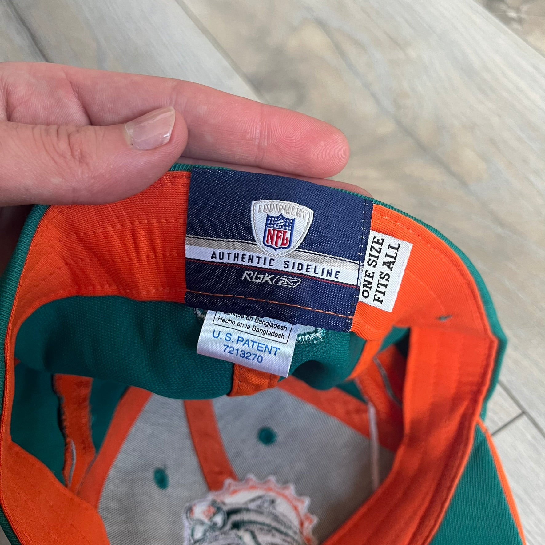 Reebok NFL Miami Dolphins Sideline Baseball Cap is a turquoise baseball cap featuring embroidered Miami Dolphins branding on the front. It includes orange and white accent detailing across the design. The side is finished with a white embroidered Reebok logo, while the back and inside features NFL Equipment tags. The construction uses cotton with a structured fit and a classic adjustable design.