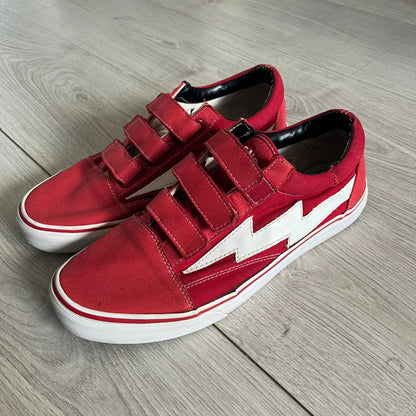 Revenge x Storm Red Strap Sneakers designed by Ian Connor. Featuring a red upper with white leather lightning bolts replacing the traditional Vans stripe. Finished with Velcro strap closures, white midsoles, black outsoles, and the signature “Revenge x Storm” branding in red on the sole.