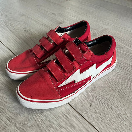 Revenge x Storm Red Strap Sneakers designed by Ian Connor. Featuring a red upper with white leather lightning bolts replacing the traditional Vans stripe. Finished with Velcro strap closures, white midsoles, black outsoles, and the signature “Revenge x Storm” branding in red on the sole.
