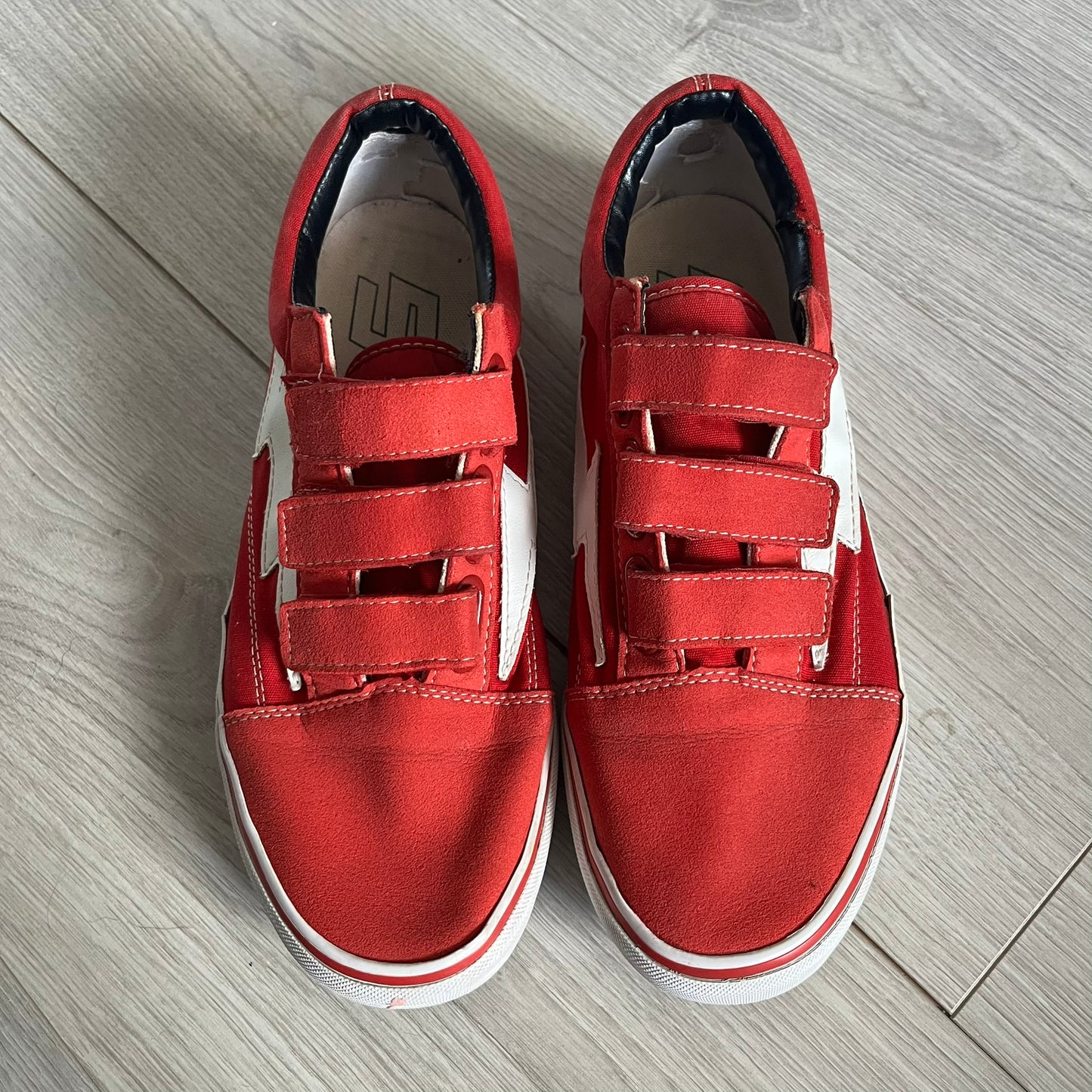 Revenge x Storm Red Strap Sneakers designed by Ian Connor. Featuring a red upper with white leather lightning bolts replacing the traditional Vans stripe. Finished with Velcro strap closures, white midsoles, black outsoles, and the signature “Revenge x Storm” branding in red on the sole.