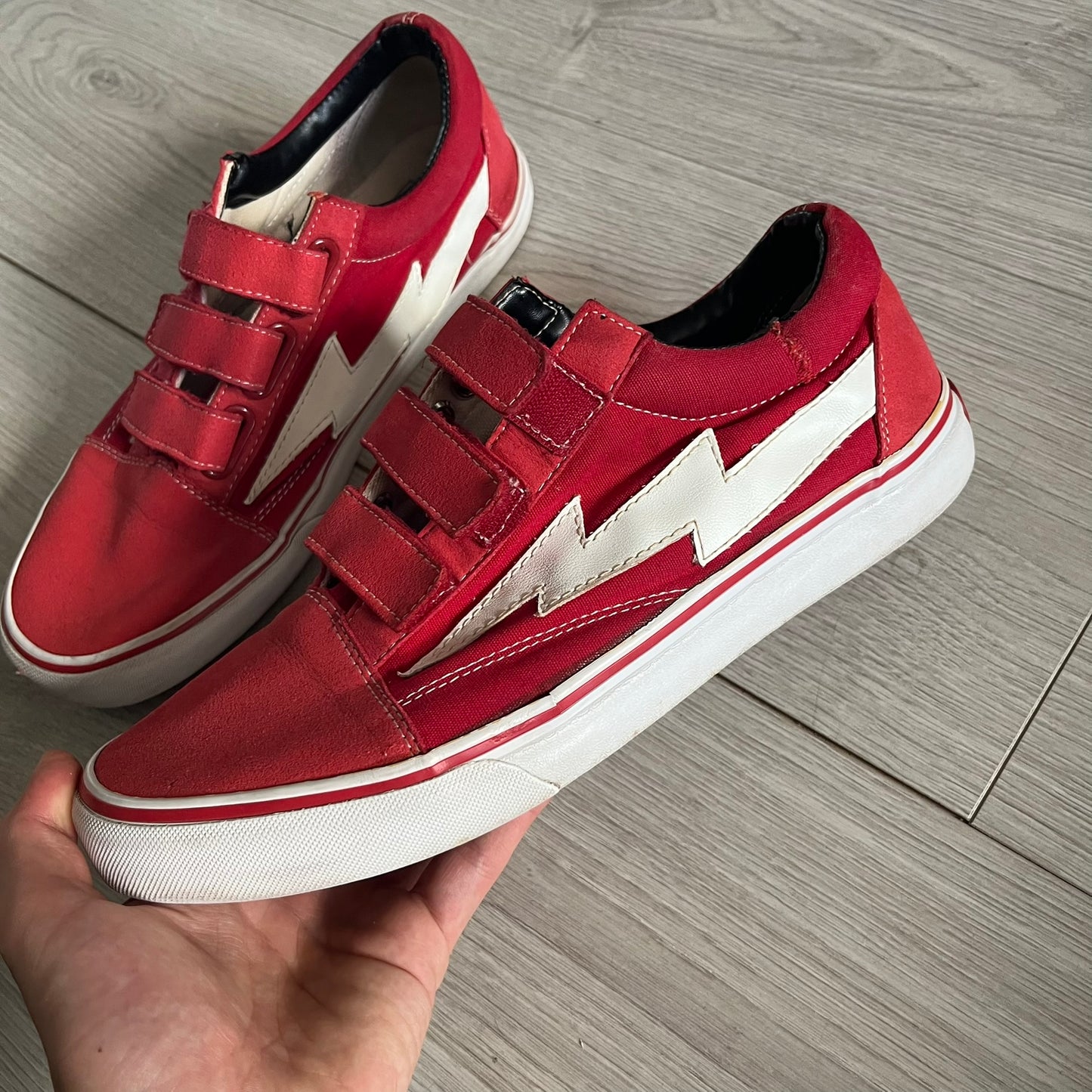 Revenge x Storm Red Strap Sneakers designed by Ian Connor. Featuring a red upper with white leather lightning bolts replacing the traditional Vans stripe. Finished with Velcro strap closures, white midsoles, black outsoles, and the signature “Revenge x Storm” branding in red on the sole.