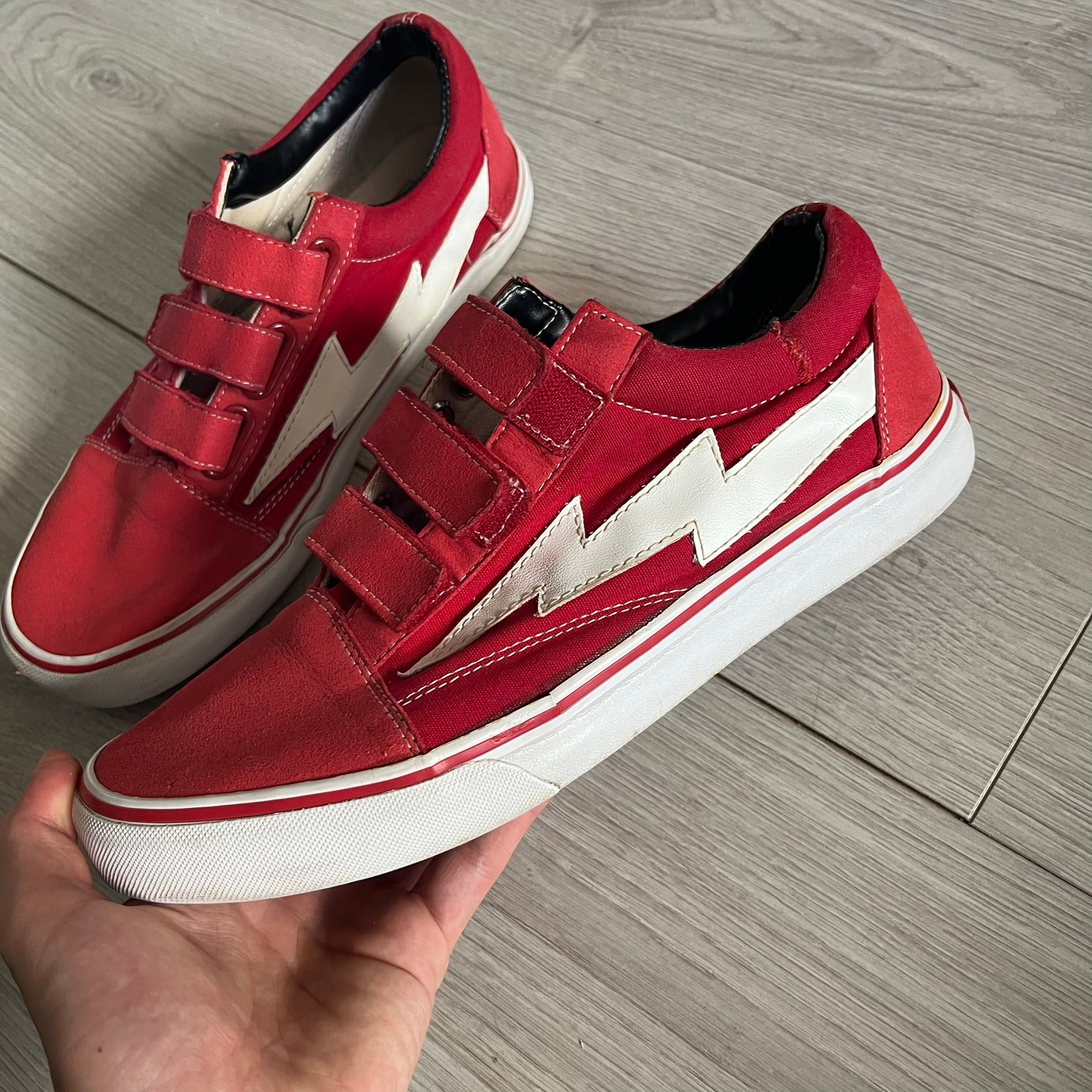 Revenge x Storm Red Strap Sneakers designed by Ian Connor. Featuring a red upper with white leather lightning bolts replacing the traditional Vans stripe. Finished with Velcro strap closures, white midsoles, black outsoles, and the signature “Revenge x Storm” branding in red on the sole.