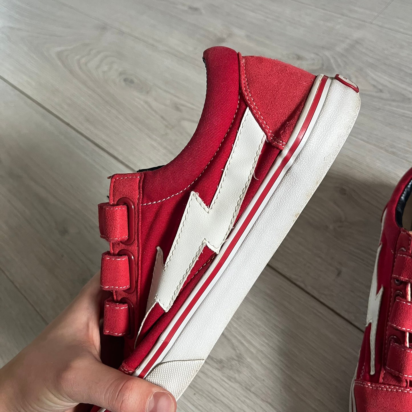 Revenge x Storm Red Strap Sneakers designed by Ian Connor. Featuring a red upper with white leather lightning bolts replacing the traditional Vans stripe. Finished with Velcro strap closures, white midsoles, black outsoles, and the signature “Revenge x Storm” branding in red on the sole.