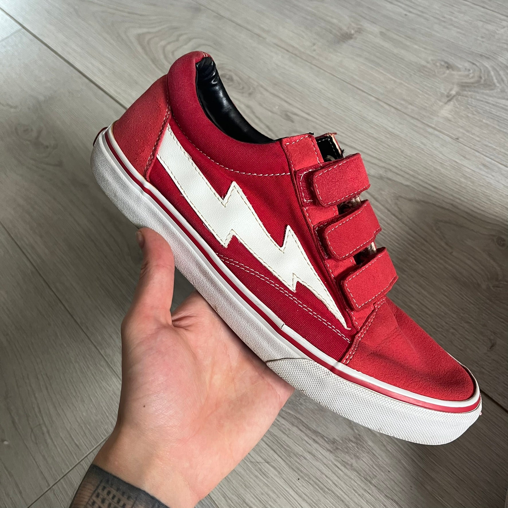 Revenge x Storm Red Strap Sneakers designed by Ian Connor. Featuring a red upper with white leather lightning bolts replacing the traditional Vans stripe. Finished with Velcro strap closures, white midsoles, black outsoles, and the signature “Revenge x Storm” branding in red on the sole.