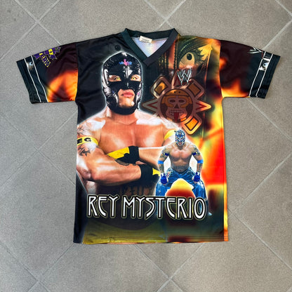Rey Mysterio 2007 Kalciomania AOP Tee features an all-over print design with red, black, and white graphics throughout. The front includes a Rey Mysterio graphic with “Rey Mysterio” text, WWE logos, and additional printed details. The back features Rey Mysterio wearing a white mask with his signature and “619” graphic. The sleeves include a WWE logo and Rey Mysterio branding. Finished with a Kalciomania licensed tag.
