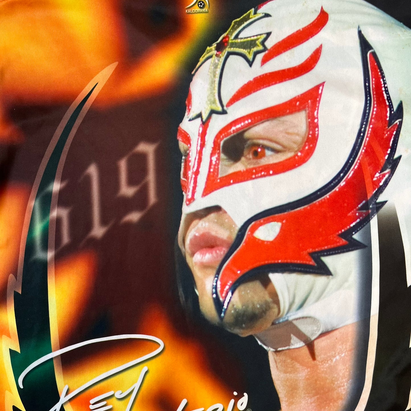 Rey Mysterio 2007 Kalciomania AOP Tee features an all-over print design with red, black, and white graphics throughout. The front includes a Rey Mysterio graphic with “Rey Mysterio” text, WWE logos, and additional printed details. The back features Rey Mysterio wearing a white mask with his signature and “619” graphic. The sleeves include a WWE logo and Rey Mysterio branding. Finished with a Kalciomania licensed tag.
