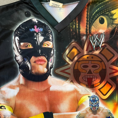 Rey Mysterio 2007 Kalciomania AOP Tee features an all-over print design with red, black, and white graphics throughout. The front includes a Rey Mysterio graphic with “Rey Mysterio” text, WWE logos, and additional printed details. The back features Rey Mysterio wearing a white mask with his signature and “619” graphic. The sleeves include a WWE logo and Rey Mysterio branding. Finished with a Kalciomania licensed tag.
