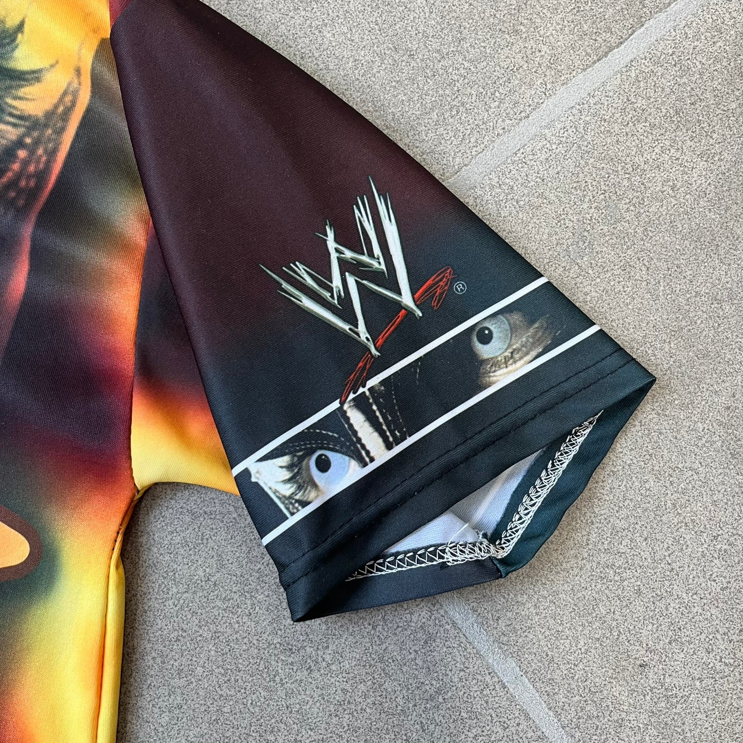 Rey Mysterio 2007 Kalciomania AOP Tee features an all-over print design with red, black, and white graphics throughout. The front includes a Rey Mysterio graphic with “Rey Mysterio” text, WWE logos, and additional printed details. The back features Rey Mysterio wearing a white mask with his signature and “619” graphic. The sleeves include a WWE logo and Rey Mysterio branding. Finished with a Kalciomania licensed tag.

