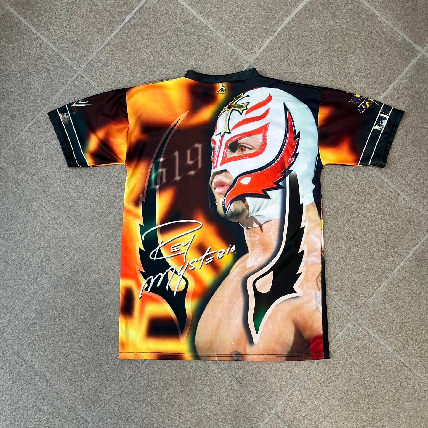 Rey Mysterio 2007 Kalciomania AOP Tee features an all-over print design with red, black, and white graphics throughout. The front includes a Rey Mysterio graphic with “Rey Mysterio” text, WWE logos, and additional printed details. The back features Rey Mysterio wearing a white mask with his signature and “619” graphic. The sleeves include a WWE logo and Rey Mysterio branding. Finished with a Kalciomania licensed tag.
