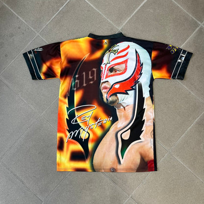 Rey Mysterio 2007 Kalciomania AOP Tee features an all-over print design with red, black, and white graphics throughout. The front includes a Rey Mysterio graphic with “Rey Mysterio” text, WWE logos, and additional printed details. The back features Rey Mysterio wearing a white mask with his signature and “619” graphic. The sleeves include a WWE logo and Rey Mysterio branding. Finished with a Kalciomania licensed tag.
