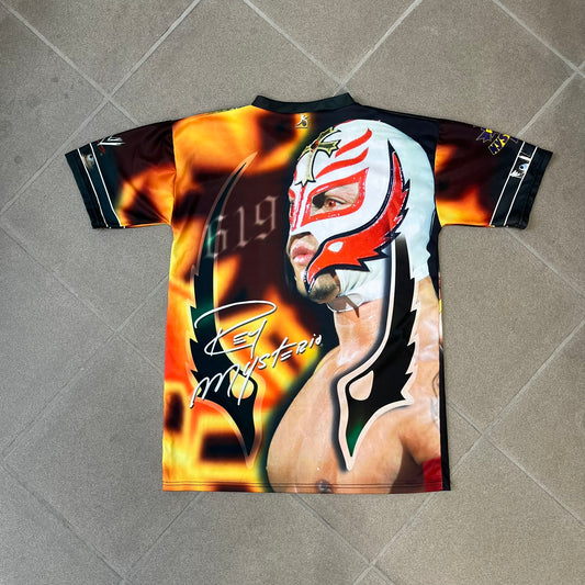 Rey Mysterio 2007 Kalciomania AOP Tee features an all-over print design with red, black, and white graphics throughout. The front includes a Rey Mysterio graphic with “Rey Mysterio” text, WWE logos, and additional printed details. The back features Rey Mysterio wearing a white mask with his signature and “619” graphic. The sleeves include a WWE logo and Rey Mysterio branding. Finished with a Kalciomania licensed tag.
