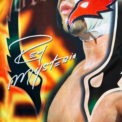 Rey Mysterio 2007 Kalciomania AOP Tee features an all-over print design with red, black, and white graphics throughout. The front includes a Rey Mysterio graphic with “Rey Mysterio” text, WWE logos, and additional printed details. The back features Rey Mysterio wearing a white mask with his signature and “619” graphic. The sleeves include a WWE logo and Rey Mysterio branding. Finished with a Kalciomania licensed tag.

