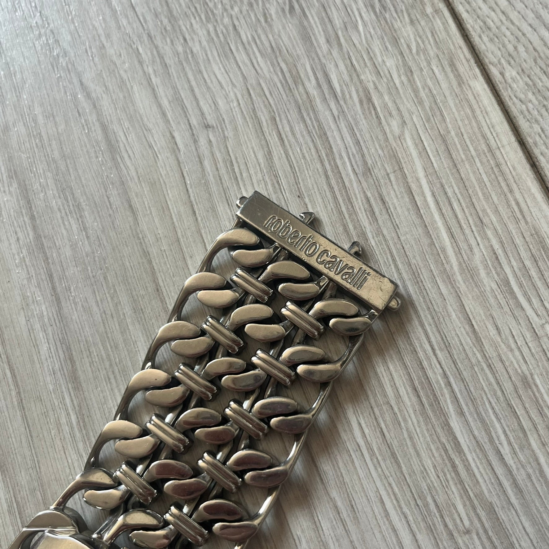 Roberto Cavalli 2000s Double Chain Steel Analog Watch is a stainless steel watch with a double-chain bracelet and a clip-style clasp. The dial has a pearlescent finish that changes appearance depending on the light. It features two asymmetrical silver hands and a stainless steel case. The watch is rated for 3 ATM water resistance.