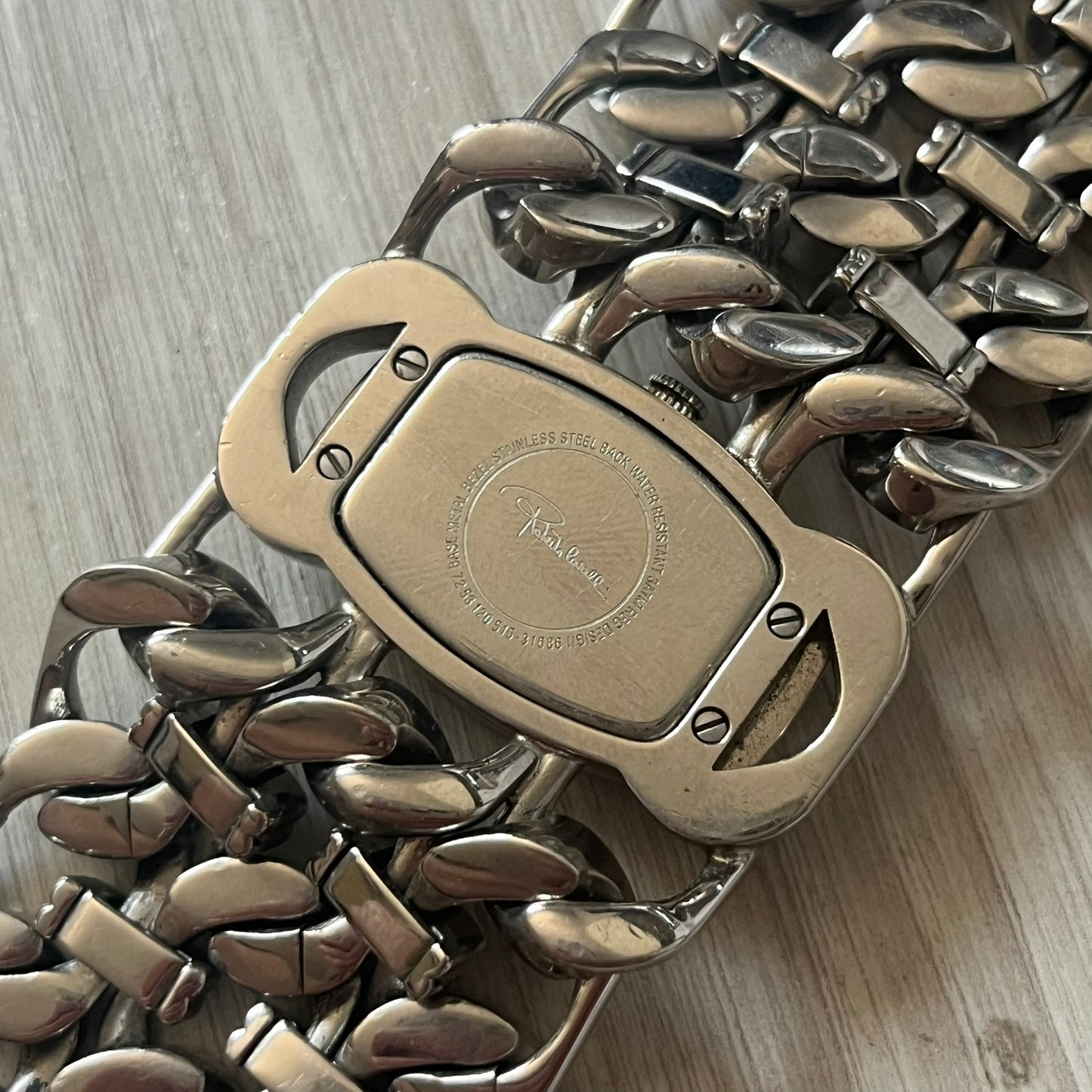 Roberto Cavalli 2000s Double Chain Steel Analog Watch is a stainless steel watch with a double-chain bracelet and a clip-style clasp. The dial has a pearlescent finish that changes appearance depending on the light. It features two asymmetrical silver hands and a stainless steel case. The watch is rated for 3 ATM water resistance.