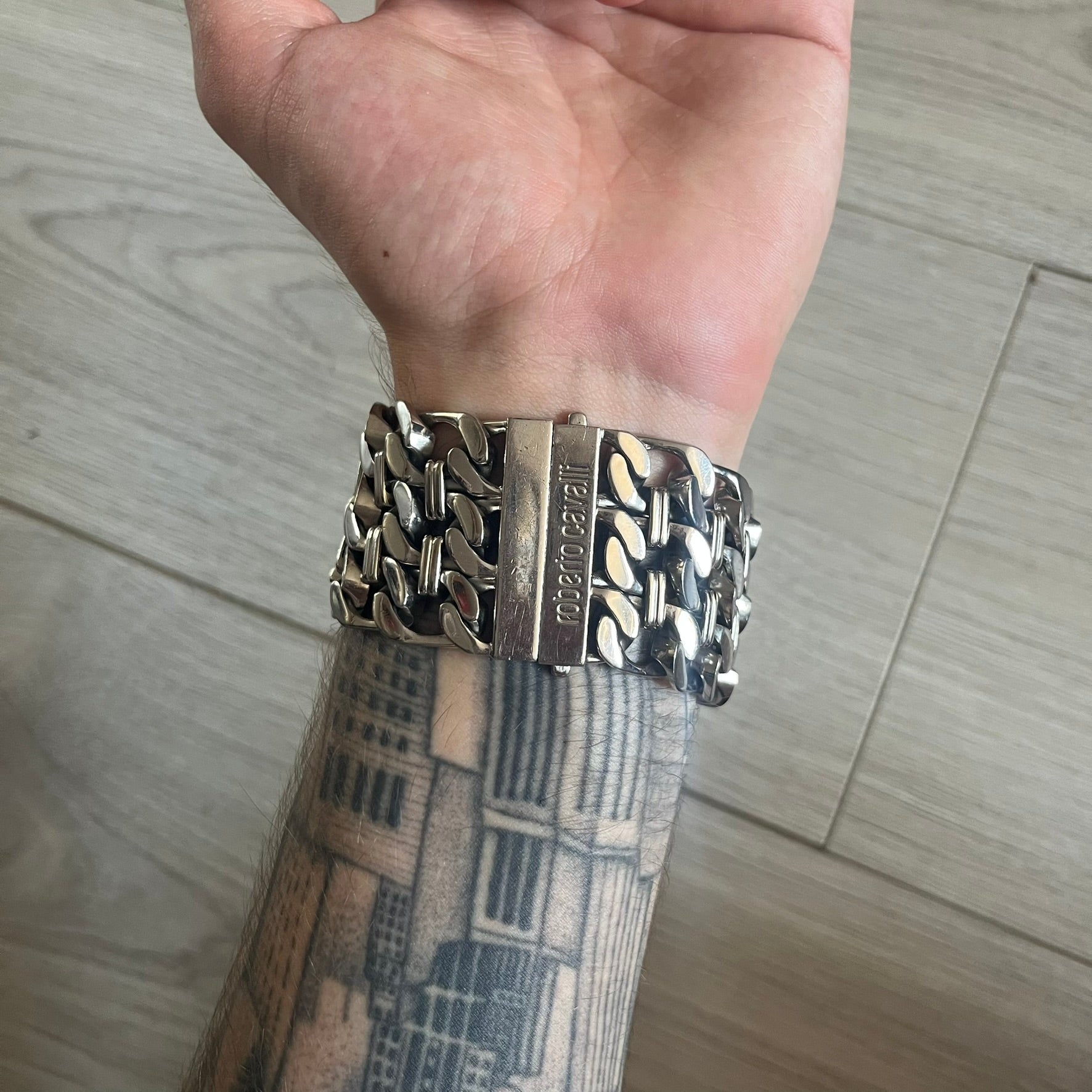 Roberto Cavalli 2000s Double Chain Steel Analog Watch is a stainless steel watch with a double-chain bracelet and a clip-style clasp. The dial has a pearlescent finish that changes appearance depending on the light. It features two asymmetrical silver hands and a stainless steel case. The watch is rated for 3 ATM water resistance.