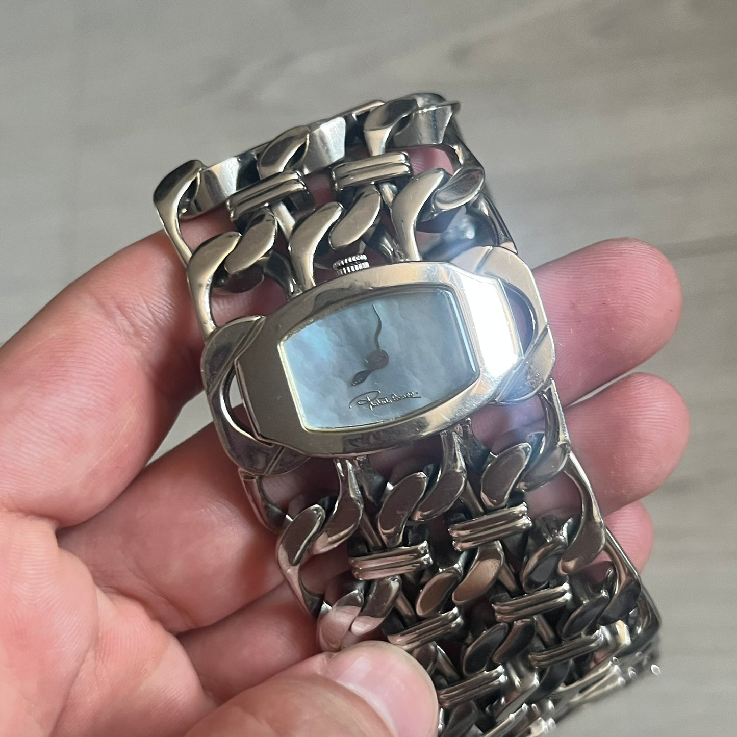 Roberto Cavalli 2000s Double Chain Steel Analog Watch is a stainless steel watch with a double-chain bracelet and a clip-style clasp. The dial has a pearlescent finish that changes appearance depending on the light. It features two asymmetrical silver hands and a stainless steel case. The watch is rated for 3 ATM water resistance.