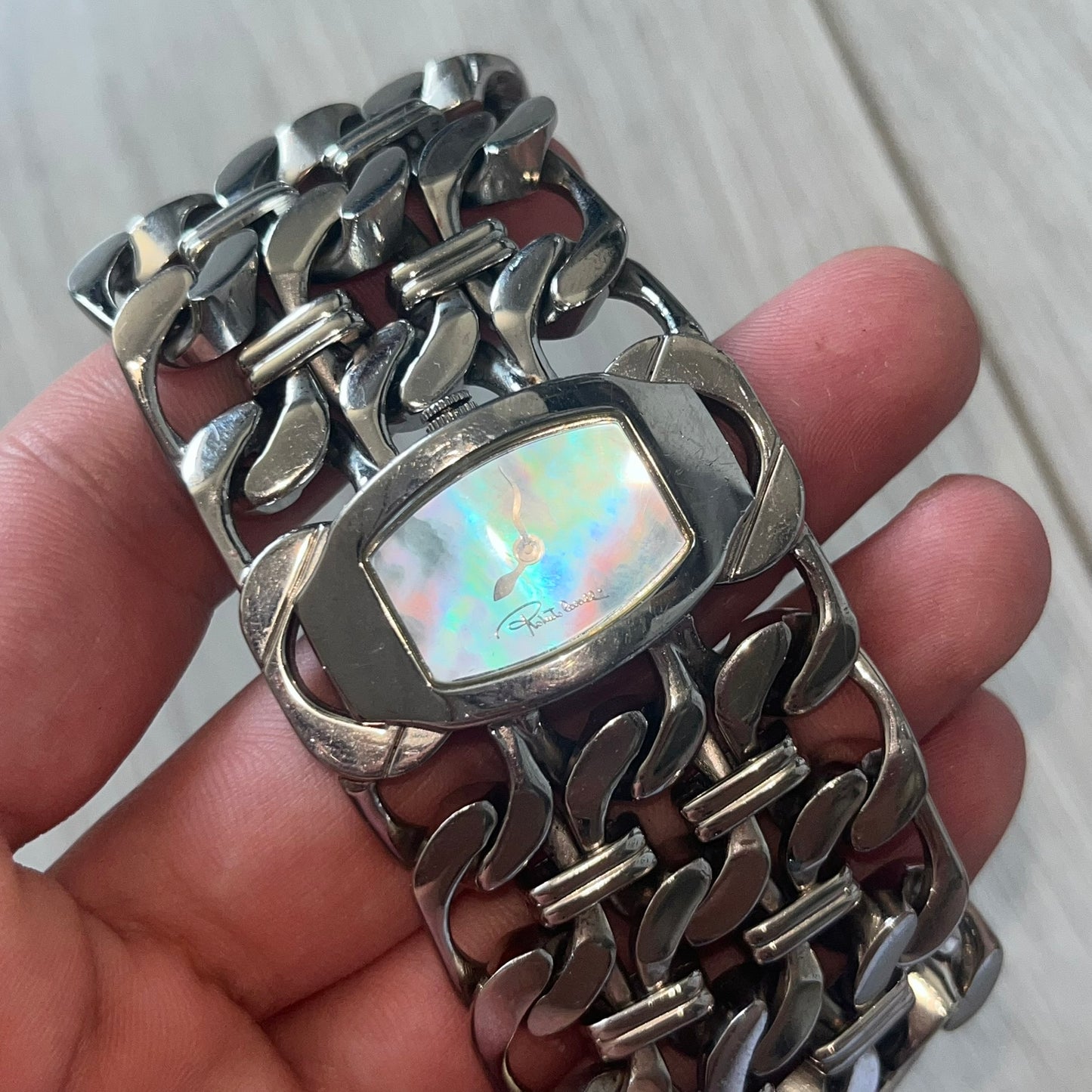 Roberto Cavalli 2000s Double Chain Steel Analog Watch is a stainless steel watch with a double-chain bracelet and a clip-style clasp. The dial has a pearlescent finish that changes appearance depending on the light. It features two asymmetrical silver hands and a stainless steel case. The watch is rated for 3 ATM water resistance.