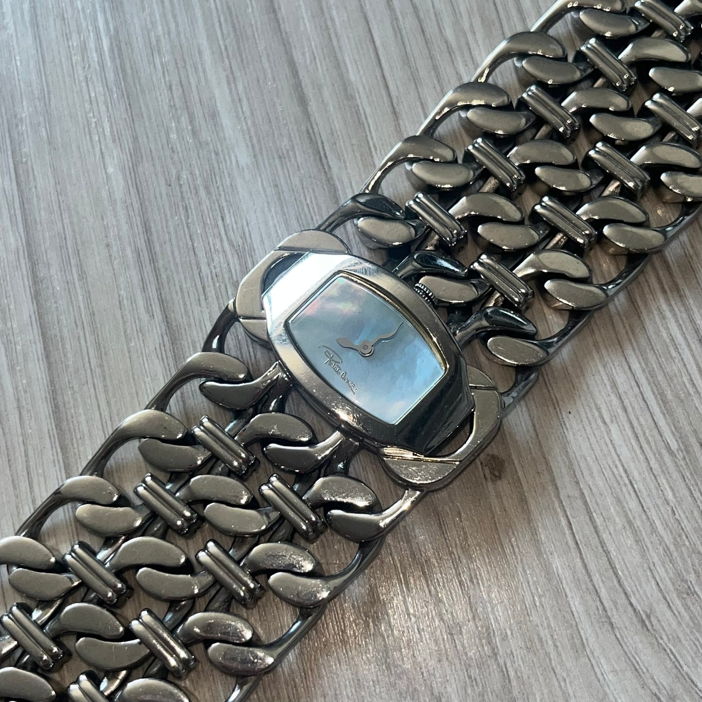 Roberto Cavalli 2000s Double Chain Steel Analog Watch is a stainless steel watch with a double-chain bracelet and a clip-style clasp. The dial has a pearlescent finish that changes appearance depending on the light. It features two asymmetrical silver hands and a stainless steel case. The watch is rated for 3 ATM water resistance.