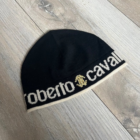 Roberto Cavalli Gold Logo Beanie is a black knit beanie made in Italy from 100% wool. The front features a raised gold Cavalli emblem, with “Roberto Cavalli” text in white integrated into the knit on both sides of the logo. The beanie has a thin wool construction suitable for lightweight wear.