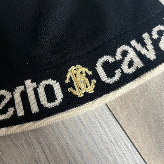 Roberto Cavalli Gold Logo Beanie is a black knit beanie made in Italy from 100% wool. The front features a raised gold Cavalli emblem, with “Roberto Cavalli” text in white integrated into the knit on both sides of the logo. The beanie has a thin wool construction suitable for lightweight wear.