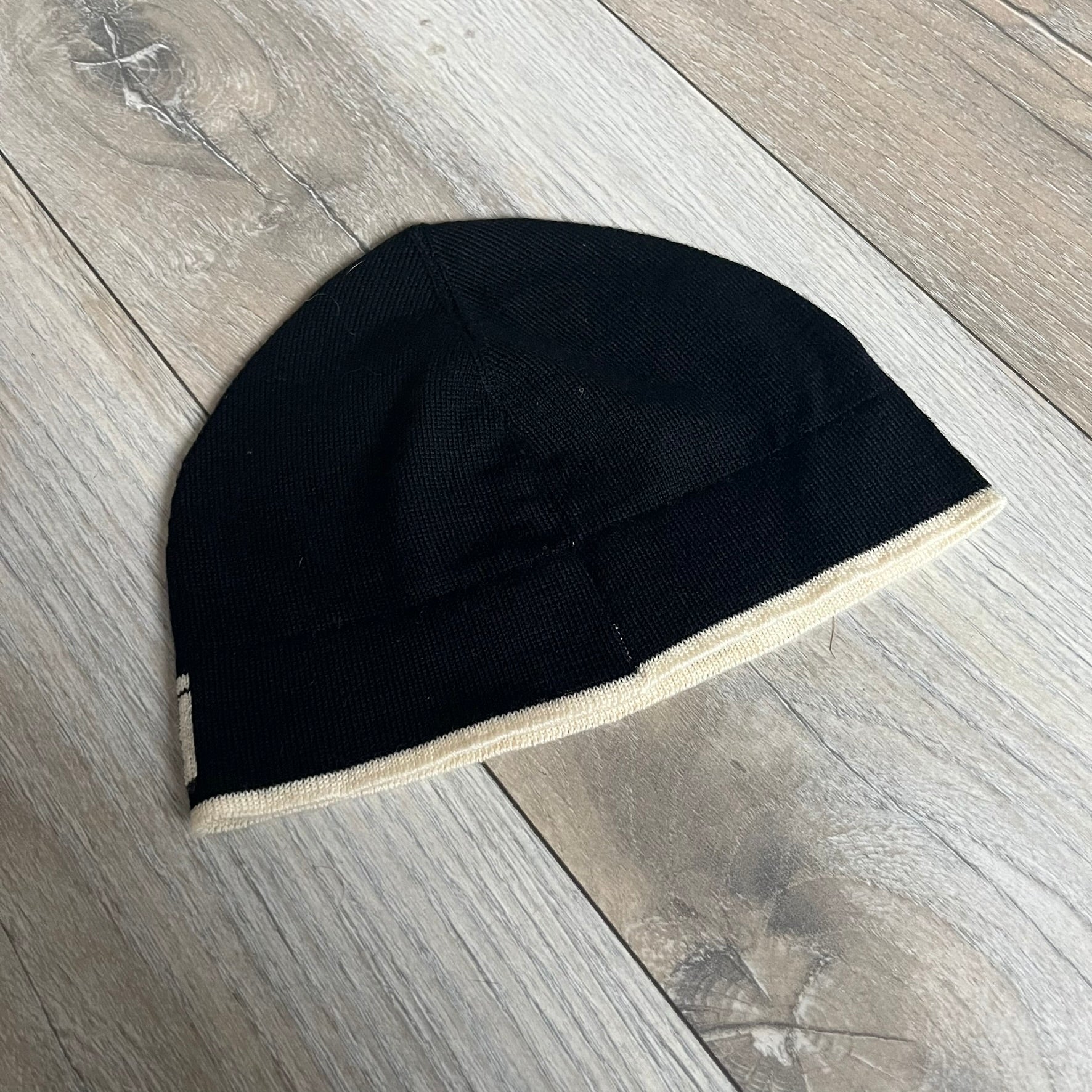Roberto Cavalli Gold Logo Beanie is a black knit beanie made in Italy from 100% wool. The front features a raised gold Cavalli emblem, with “Roberto Cavalli” text in white integrated into the knit on both sides of the logo. The beanie has a thin wool construction suitable for lightweight wear.