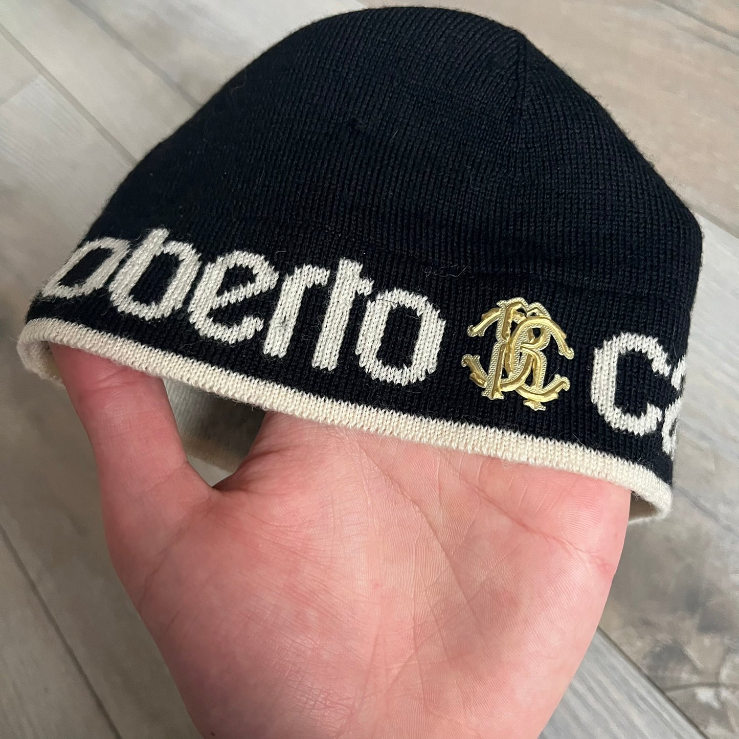 Roberto Cavalli Gold Logo Beanie is a black knit beanie made in Italy from 100% wool. The front features a raised gold Cavalli emblem, with “Roberto Cavalli” text in white integrated into the knit on both sides of the logo. The beanie has a thin wool construction suitable for lightweight wear.