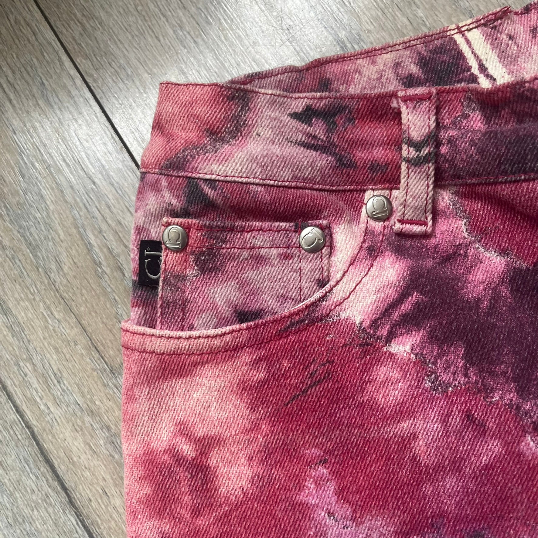 Roberto Cavalli Pink Floral 2000’s Jeans are made in Italy from 98% cotton and 2% elastane. The jeans feature an all-over floral print in multiple shades of pink, ranging from light to dark, with a hand-painted effect across the fabric. Details include branded buttons, a leather logo tab at the back, and classic five-pocket construction with two front and two back pockets.