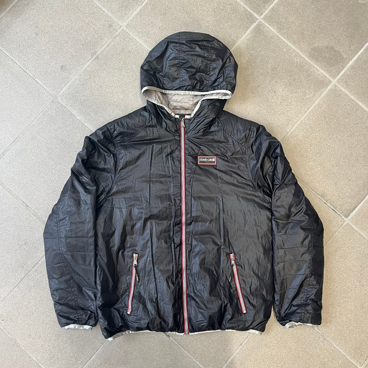 Roberto Cavalli Sport 2010's Black Lightweight Jacket is a lightweight hooded jacket in black with a gray interior, featuring two front pockets and star logo zipper pulls. A rubber Roberto Cavalli Sport logo patch sits on the chest, with a smaller matching patch on the upper back.