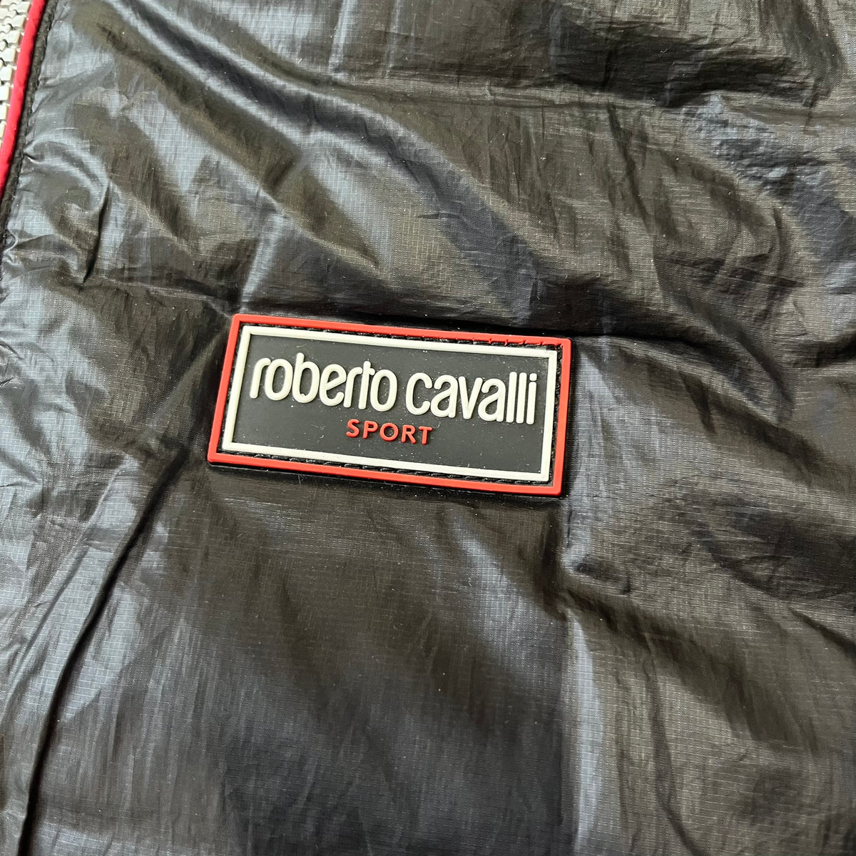 Roberto Cavalli Sport 2010's Black Lightweight Jacket is a lightweight hooded jacket in black with a gray interior, featuring two front pockets and star logo zipper pulls. A rubber Roberto Cavalli Sport logo patch sits on the chest, with a smaller matching patch on the upper back.
