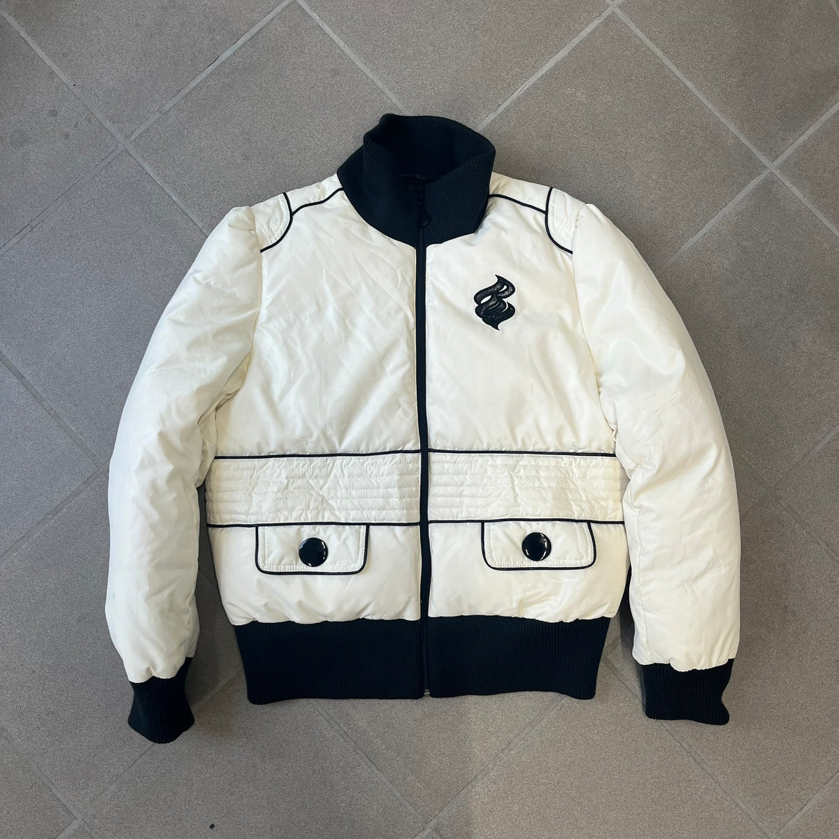 Rocawear 2000’s White Bomber Jacket is a white women’s bomber jacket with black details and a black Rocawear “R” logo on the left chest. It has two front pockets with oversized black buttons and a branded zipper, and a Rocawear logo runs across the upper back.