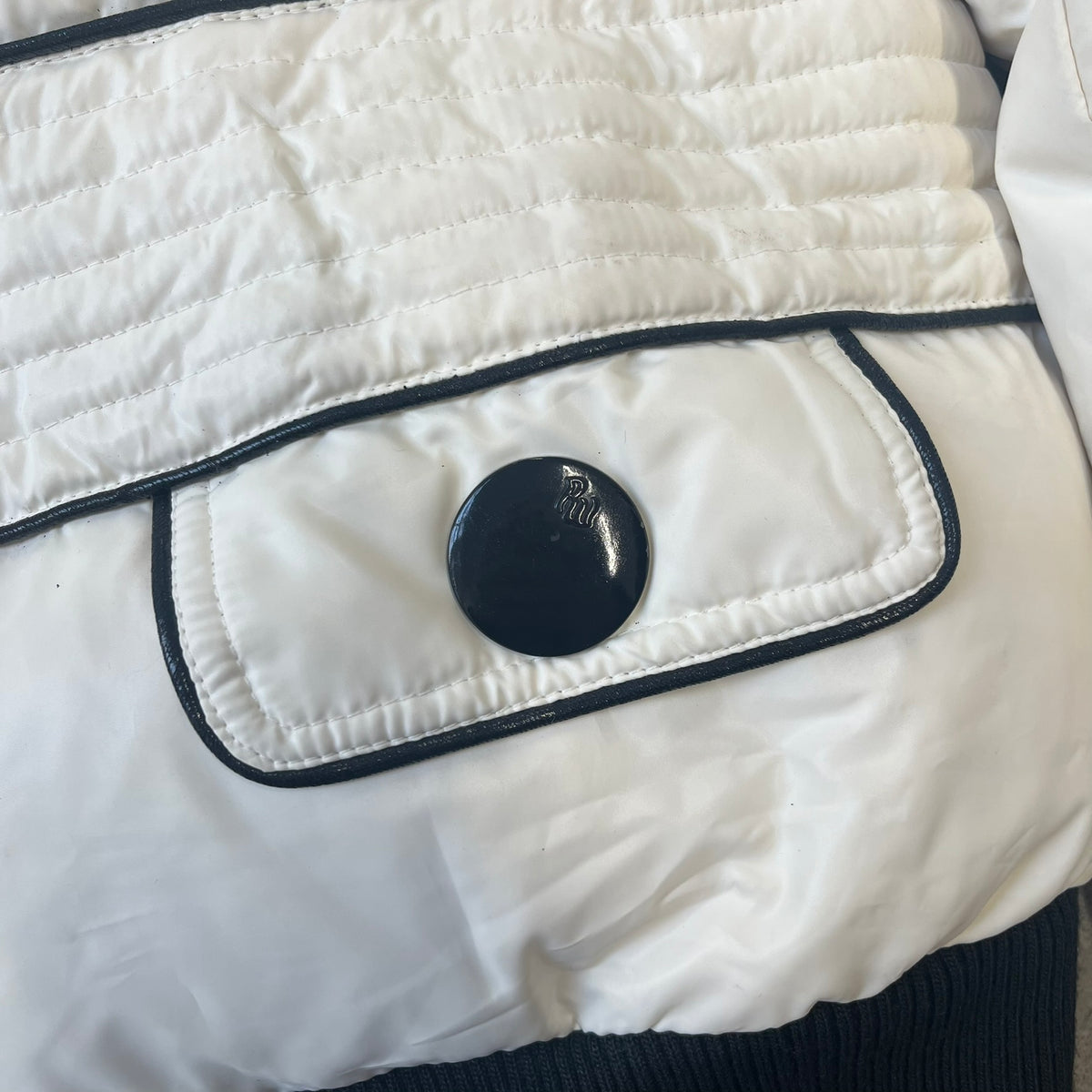 Rocawear 2000’s White Bomber Jacket is a white women’s bomber jacket with black details and a black Rocawear “R” logo on the left chest. It has two front pockets with oversized black buttons and a branded zipper, and a Rocawear logo runs across the upper back.