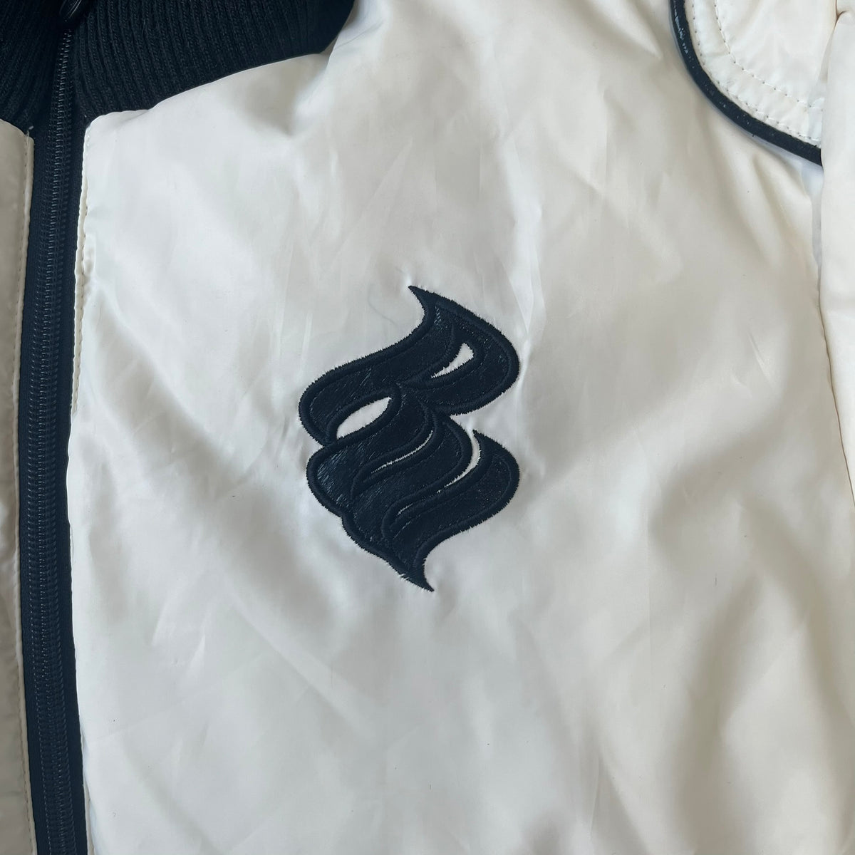 Rocawear 2000’s White Bomber Jacket is a white women’s bomber jacket with black details and a black Rocawear “R” logo on the left chest. It has two front pockets with oversized black buttons and a branded zipper, and a Rocawear logo runs across the upper back.