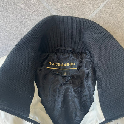 Rocawear 2000’s White Bomber Jacket is a white women’s bomber jacket with black details and a black Rocawear “R” logo on the left chest. It has two front pockets with oversized black buttons and a branded zipper, and a Rocawear logo runs across the upper back.
