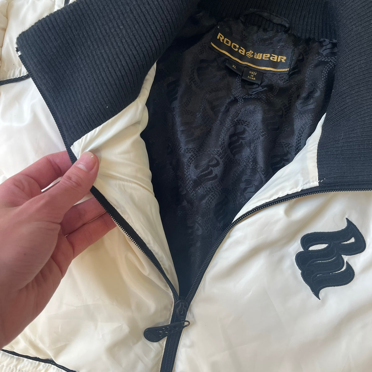 Rocawear 2000’s White Bomber Jacket is a white women’s bomber jacket with black details and a black Rocawear “R” logo on the left chest. It has two front pockets with oversized black buttons and a branded zipper, and a Rocawear logo runs across the upper back.