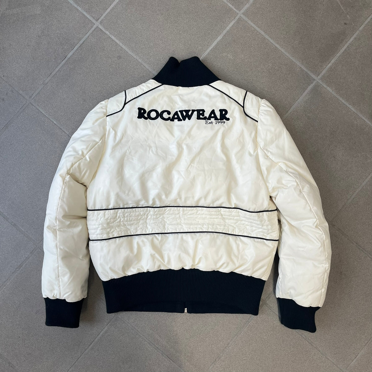 Rocawear 2000’s White Bomber Jacket is a white women’s bomber jacket with black details and a black Rocawear “R” logo on the left chest. It has two front pockets with oversized black buttons and a branded zipper, and a Rocawear logo runs across the upper back.