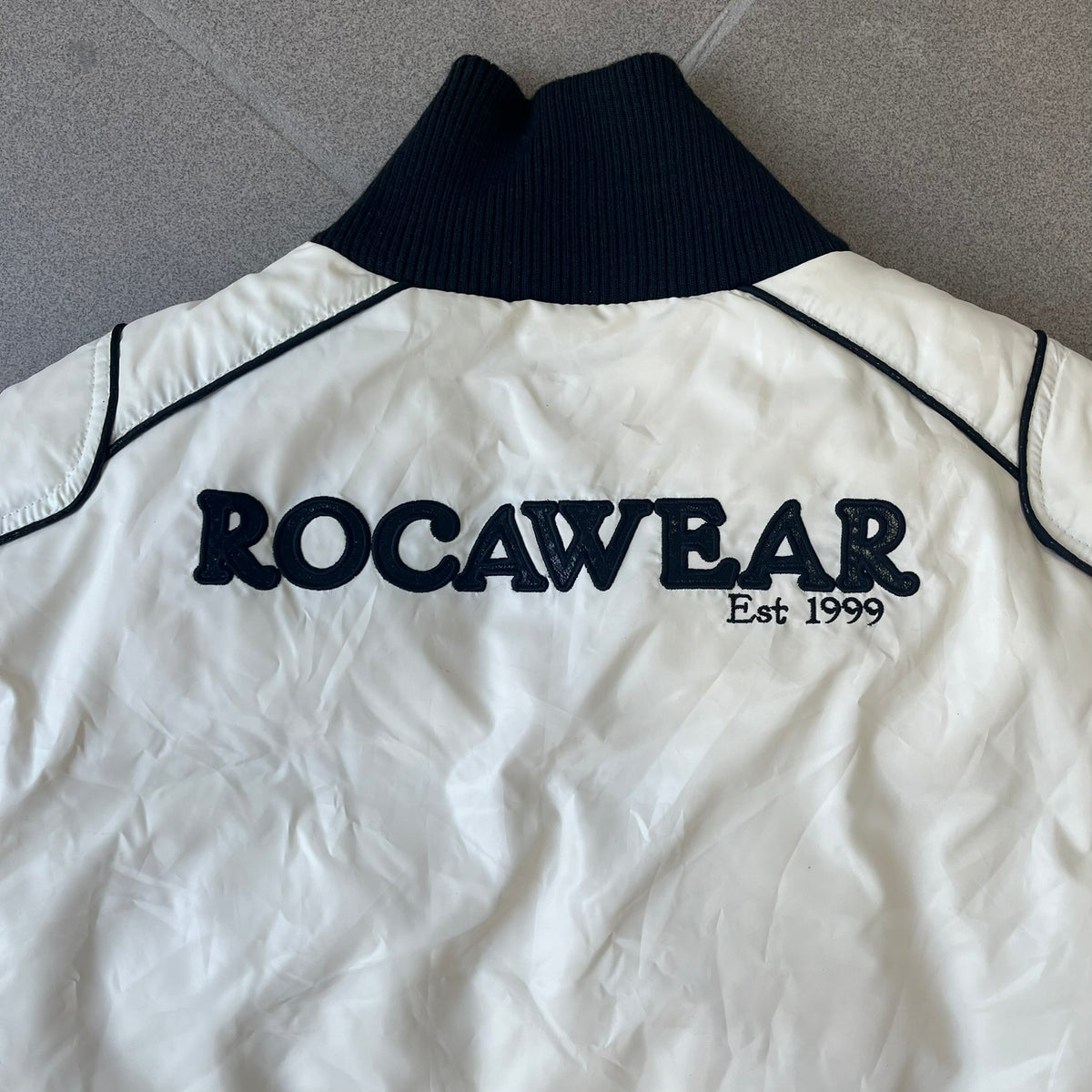 Rocawear 2000’s White Bomber Jacket is a white women’s bomber jacket with black details and a black Rocawear “R” logo on the left chest. It has two front pockets with oversized black buttons and a branded zipper, and a Rocawear logo runs across the upper back.