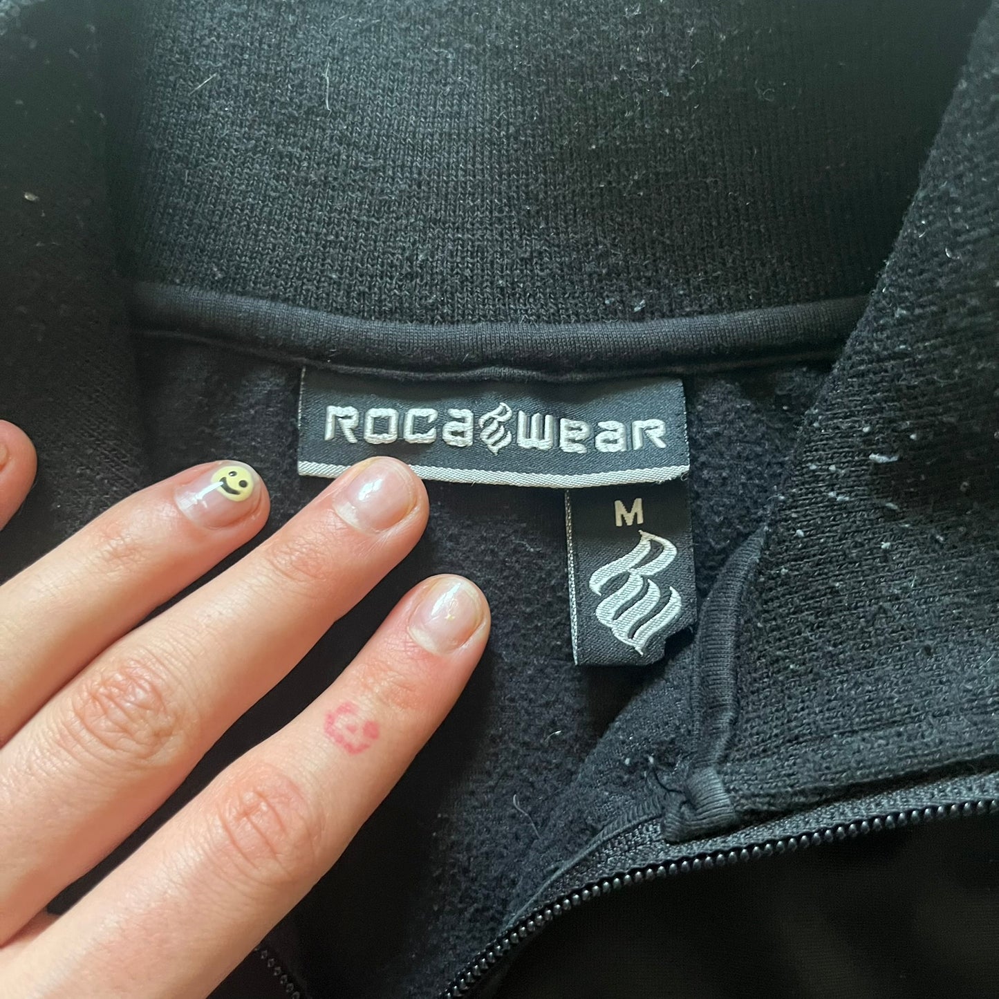 Vintage Rocawear black track jacket featuring a small embroidered chest logo and big white branding across the back. Silver zipper and wrist emblem add refined details to this early-2000s streetwear classic.