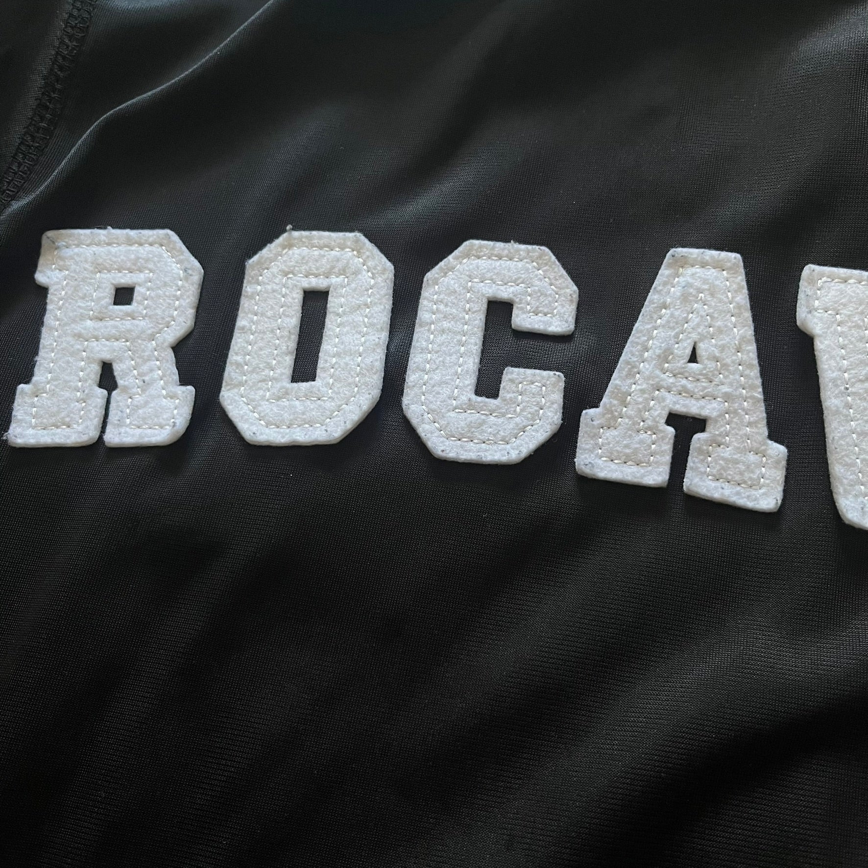 Vintage Rocawear black track jacket featuring a small embroidered chest logo and big white branding across the back. Silver zipper and wrist emblem add refined details to this early-2000s streetwear classic.