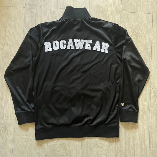 Vintage Rocawear black track jacket featuring a small embroidered chest logo and big white branding across the back. Silver zipper and wrist emblem add refined details to this early-2000s streetwear classic.