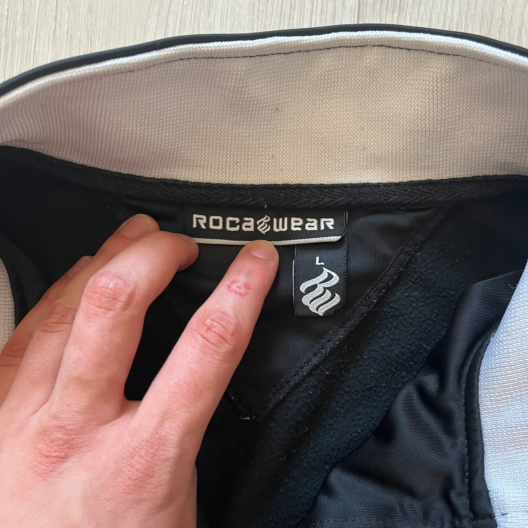 Rocawear black track jacket in size large, designed with a small embroidered logo on the chest and a large white logo across the back. A silver emblem near the wrist and a detailed silver zipper add distinctive touches. A sleek early 2000s streetwear piece.