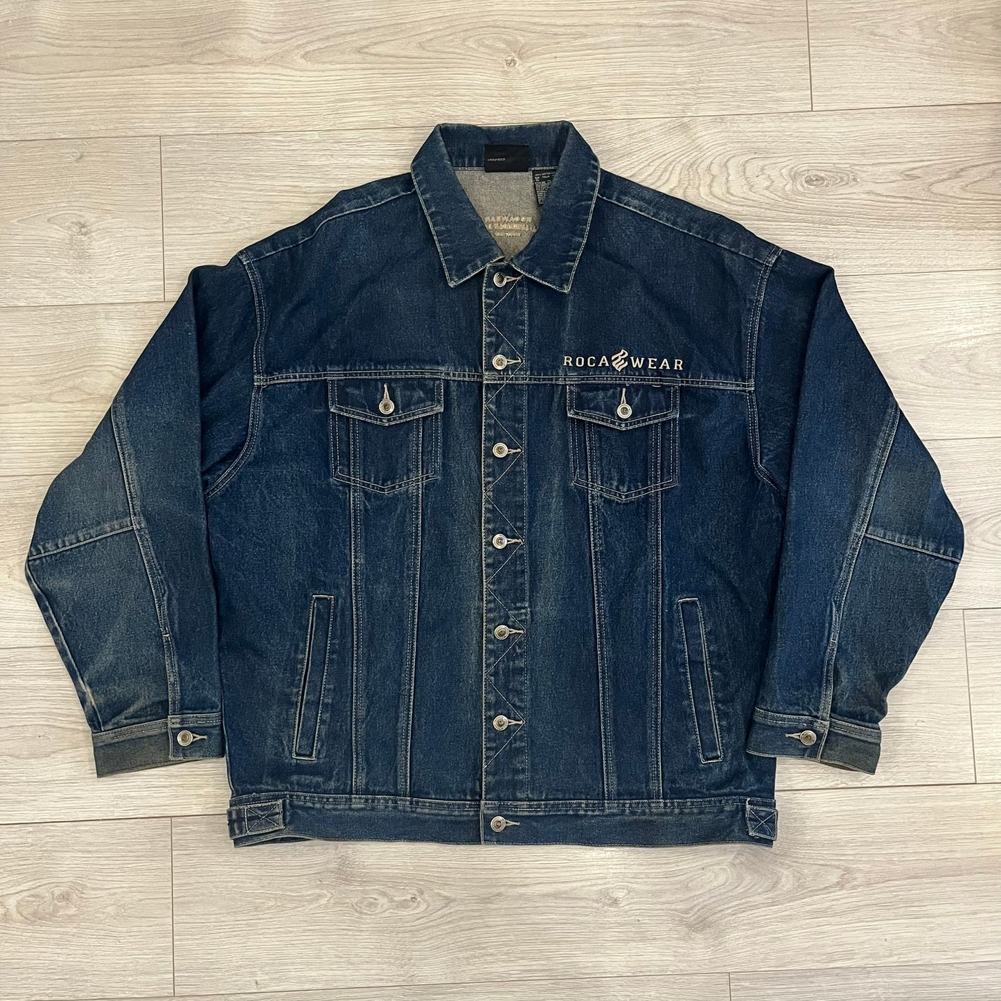 Rocawear blue denim jacket from the early 2000s, made from 100% cotton. Features an embroidered Rocawear logo on the right chest above the pocket, two buttoned chest pockets and standard side pockets. Finished with branded Rocawear metal buttons, a leather tab with metal R logo at the lower back, and “Rocawear All American Denim Industries” embroidery placed under the collar on the back. Classic structured denim fit with durable construction.