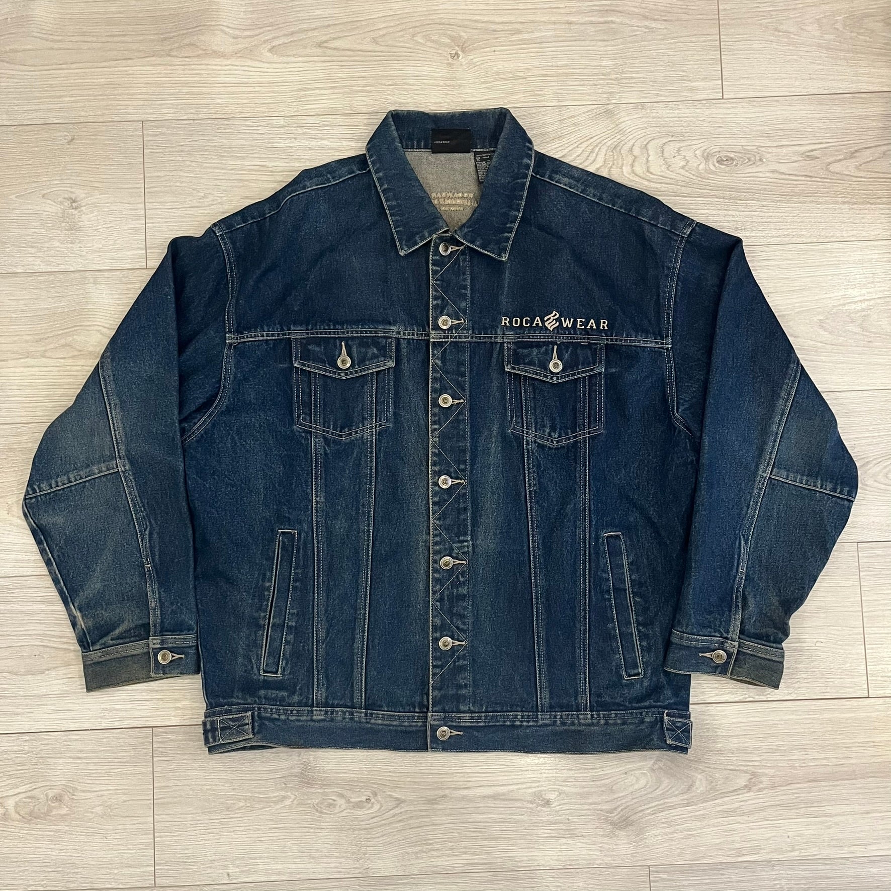 Rocawear blue denim jacket from the early 2000s, made from 100% cotton. Features an embroidered Rocawear logo on the right chest above the pocket, two buttoned chest pockets and standard side pockets. Finished with branded Rocawear metal buttons, a leather tab with metal R logo at the lower back, and “Rocawear All American Denim Industries” embroidery placed under the collar on the back. Classic structured denim fit with durable construction.