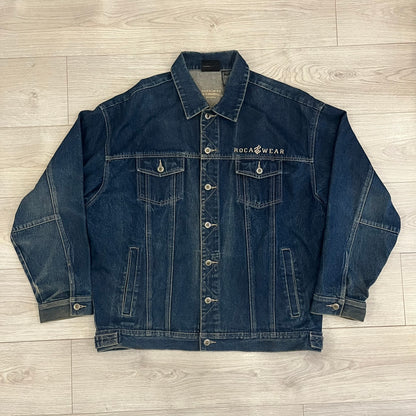 Rocawear blue denim jacket from the early 2000s, made from 100% cotton. Features an embroidered Rocawear logo on the right chest above the pocket, two buttoned chest pockets and standard side pockets. Finished with branded Rocawear metal buttons, a leather tab with metal R logo at the lower back, and “Rocawear All American Denim Industries” embroidery placed under the collar on the back. Classic structured denim fit with durable construction.