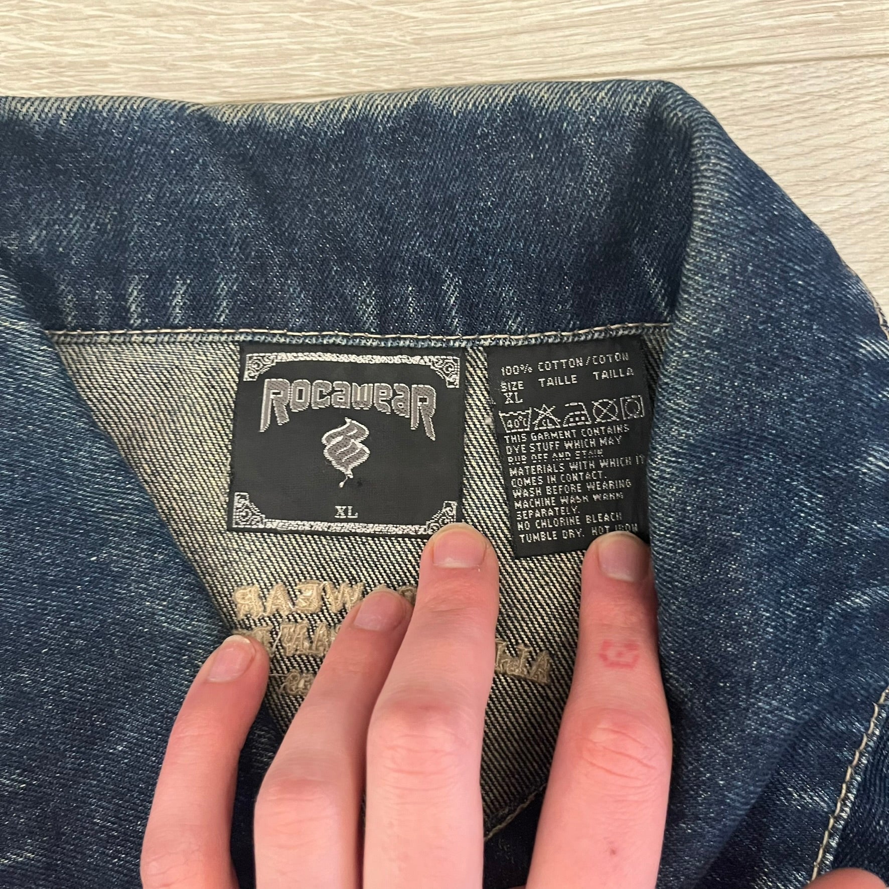 Rocawear blue denim jacket from the early 2000s, made from 100% cotton. Features an embroidered Rocawear logo on the right chest above the pocket, two buttoned chest pockets and standard side pockets. Finished with branded Rocawear metal buttons, a leather tab with metal R logo at the lower back, and “Rocawear All American Denim Industries” embroidery placed under the collar on the back. Classic structured denim fit with durable construction.