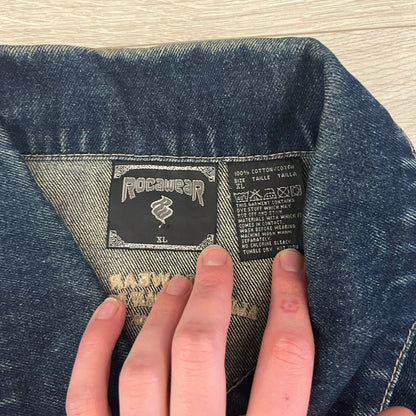 Rocawear blue denim jacket from the early 2000s, made from 100% cotton. Features an embroidered Rocawear logo on the right chest above the pocket, two buttoned chest pockets and standard side pockets. Finished with branded Rocawear metal buttons, a leather tab with metal R logo at the lower back, and “Rocawear All American Denim Industries” embroidery placed under the collar on the back. Classic structured denim fit with durable construction.