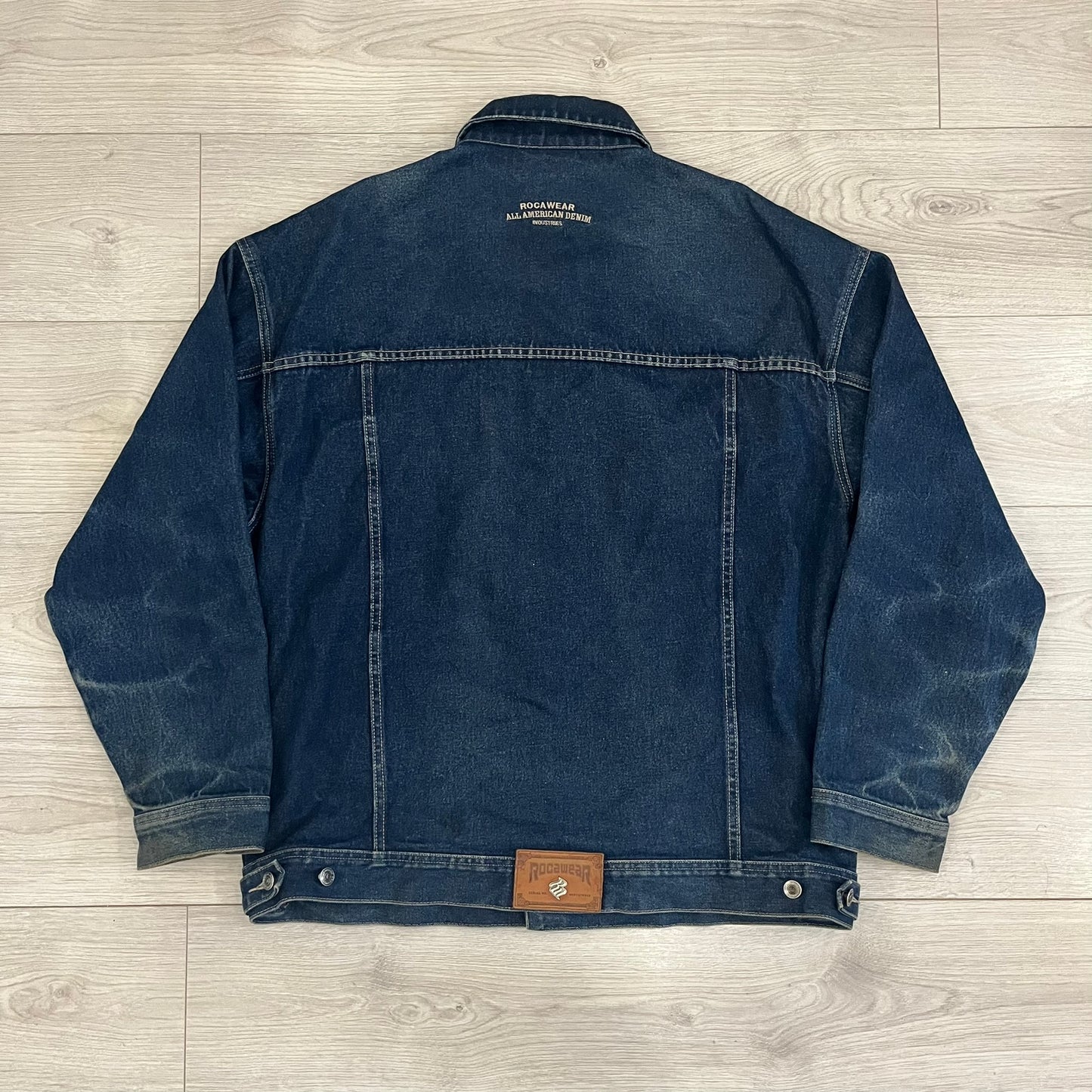 Rocawear blue denim jacket from the early 2000s, made from 100% cotton. Features an embroidered Rocawear logo on the right chest above the pocket, two buttoned chest pockets and standard side pockets. Finished with branded Rocawear metal buttons, a leather tab with metal R logo at the lower back, and “Rocawear All American Denim Industries” embroidery placed under the collar on the back. Classic structured denim fit with durable construction.